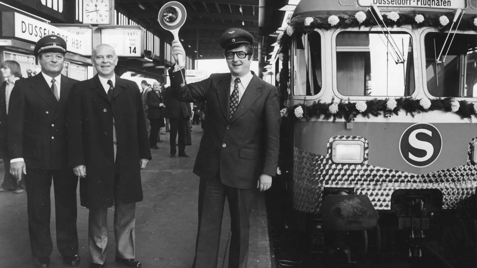 Historische Aufnahme einer S-Bahn am Düsseldorfer Hauptbahnhof. Vor dem Zug steht das Personal, der Kontrolleur hält eine Kelle in der Hand.